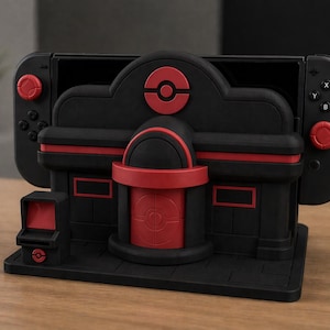 Puede incluir: Soporte negro y rojo para consola de juegos, diseñado para parecerse a un edificio con un logotipo circular en la parte superior. El soporte tiene una ranura para sujetar la consola y los mandos. El soporte es de plástico.