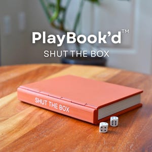 Op de afbeelding: Een oranje "Shut the Box" spel met witte dobbelstenen op een houten tafel. De speldoos is rechthoekig met witte tekst. Het spel heet "PlayBook'd". De dobbelstenen zijn wit met zwarte stippen.