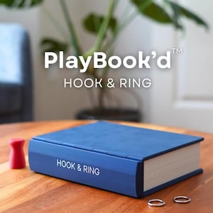 Op de afbeelding: Een blauwe PlayBook'd Hook & Ring-spelset op een houten tafel. De set bevat een blauwe, boekvormige doos, twee zilveren ringen en een klein rood object. De tekst "PlayBook'd" en "HOOK & RING" is zichtbaar op de doos.