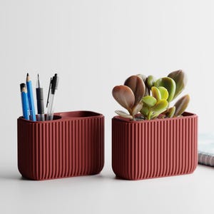 Könnte beinhalten: Zwei passende Schreibtisch-Organizer in Terrakotta-Farbe. Einer enthält Stifte und Bleistifte, der andere eine kleine Sukkulentenpflanze. Beide haben ein geripptes Design. Ein Notizbuch ist im Hintergrund teilweise sichtbar.
