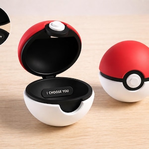 Puede incluir: Dos contenedores rojos y blancos con temática de Poké Ball, uno abierto y otro cerrado, están sobre una superficie de madera clara. El contenedor abierto revela un interior negro con el texto "I CHOOSE YOU". El contenedor cerrado tiene un botón blanco.