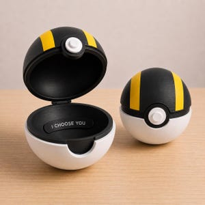 Könnte beinhalten: Zwei kugelförmige Behälter im Design von Pokéballs, mit einer schwarzen oberen Hälfte mit gelben Streifen, einer weißen unteren Hälfte und einem weißen Knopf in der Mitte. Einer ist geöffnet und zeigt den Text I CHOOSE YOU innen.