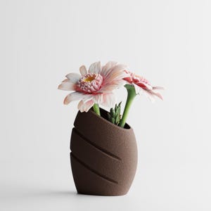 Op de afbeelding: Een bruine, getextureerde vaas met diagonale uitgesneden lijnen, met twee roze en witte gerbera's. De vaas heeft een unieke, asymmetrische vorm en de bloemen voegen een vleugje kleur toe tegen de neutrale achtergrond.