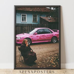 Puede incluir: Un póster enmarcado con la fotografía de una persona mayor con pañuelo y ropa oscura, sentada en una silla plegable. Al fondo, un sedán rosa brillante con calcomanías de llamas está estacionado frente a una casa azul con una valla de madera.
