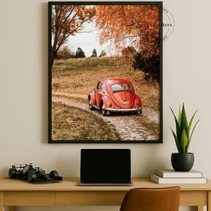 May include: A framed wall art print featuring a red vintage Volkswagen Beetle parked on a dirt path surrounded by autumn trees with orange foliage. The scene captures the rear view of the car in a rural landscape setting with a clear sky above.