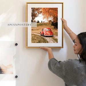 May include: A framed wall art print featuring a red vintage car on a dirt path surrounded by autumn trees with orange foliage. The print is encased in a thin gold frame with a white mat border. The text APEXXPOSTERS is visible on the left side.