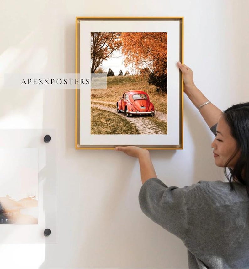 May include: A framed wall art print featuring a red vintage car on a dirt path surrounded by autumn trees with orange foliage. The print is encased in a thin gold frame with a white mat border. The text APEXXPOSTERS is visible on the left side.