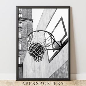 Könnte beinhalten: Ein schwarz-weiß gerahmter Wandkunstdruck, der einen Basketball zeigt, der durch ein an einem Brett befestigtes Netz fliegt. Im Hintergrund steht ein hohes Backsteingebäude neben einer Betonmauer. Unten ist der Text APEXXPOSTERS aufgedruckt.