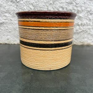 Puede incluir: Un recipiente cilíndrico de cerámica con superficie texturizada y borde marrón oscuro. El cuerpo presenta bandas horizontales de color beige, naranja, marrón y negro. La base es de color beige claro.