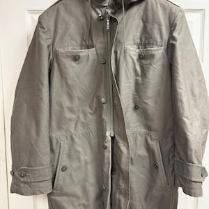 Op de afbeelding: Een lange, olijfgroene jas met capuchon, ritssluiting en knopen. De jas heeft twee voorzakken en manchetten met knopen. De jas hangt aan een zwarte hanger.