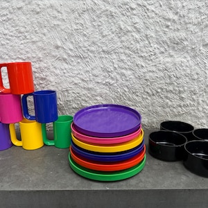 May include: A collection of colorful kitchenware including mugs, plates, and bowls. The mugs are stacked in a pyramid shape, with plates and bowls arranged nearby. The items are in various bright colors, including red, pink, blue, yellow, green, and purple.