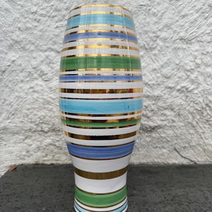 May include: A tall, cylindrical vase with horizontal stripes in shades of blue, green, white, and gold. The vase has a flared top and a rounded base. The gold stripes add a touch of elegance to the design. The vase is likely ceramic or glass.