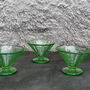Può includere: Cinque coppette per dessert in vetro verde con un design a coste. Le coppette sono disposte in fila su una superficie grigia.