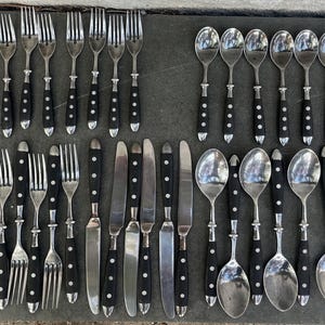 May include: A collection of stainless steel silverware with black handles. The set includes forks, knives, and spoons, arranged neatly on a dark surface. The black handles have a white dot pattern.