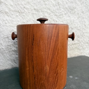Puede incluir: Una cubitera cilíndrica de madera con tapa y dos asas laterales. La cubitera es de madera de color marrón rojizo con vetas visibles y un pomo marrón oscuro en la tapa. Las asas son pequeñas y redondeadas.