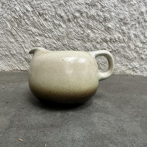May include: A ceramic creamer pitcher with a rounded body and a small handle. The pitcher features a speckled off-white glaze that transitions into a darker brown tone at the base, with a small spout for pouring liquids on the upper left side.