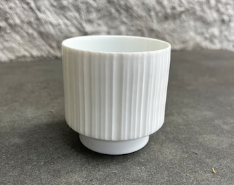 Może przedstawiać: Biała ceramiczna doniczka z żebrowanym wzorem. Doniczka ma okrągłą podstawę i cylindryczny kształt.