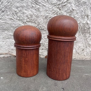 Może przedstawiać: Dwa drewniane solniczki i pieprzniczki z ciemnobrązowym wykończeniem. Solniczki są cylindryczne z zaokrąglonym wierzchołkiem i lekko rozszerzoną podstawą.