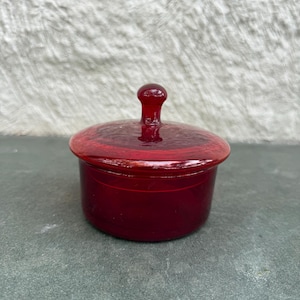 Puede incluir: Un pequeño tarro redondo de cerámica rojo rubí con tapa a juego. La tapa tiene un pequeño asa redondeada. El tarro y la tapa tienen un acabado brillante. El tarro está sobre una superficie gris.