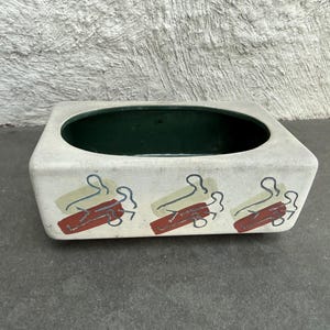 Op de afbeelding: Rechthoekige keramische plantenbak met een lichtgrijze buitenkant en een donkergroene binnenkant. De plantenbak is versierd met drie sets abstracte vormen in rood, groen en blauw. De plantenbak staat op een grijze ondergrond.