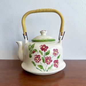 May include: Cream-colored ceramic teapot with a floral design featuring red flowers and green leaves. The teapot has a woven handle and a lid with a green rim. The spout is on the left side.