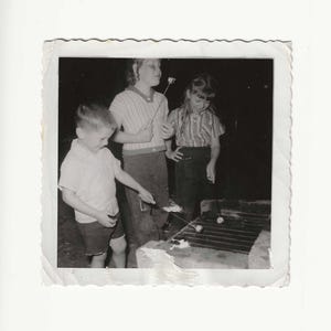 May include: Black and white vintage photograph of three children roasting marshmallows over a grill. The children are wearing casual clothing. The photo has a white border with a torn edge.