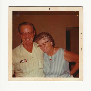 May include: Vintage photograph of a smiling man and woman. The man wears glasses and a light yellow shirt with a small emblem. The woman wears glasses, a striped blue and white top, and a necklace with a red pendant. The photo is dated 'JUN 71'.