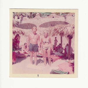 May include: Vintage photograph of a couple standing on a beach, surrounded by other people and thatched umbrellas. The woman wears a floral bikini, and the man wears dark swim trunks and a straw hat. The image has a warm, faded color palette.