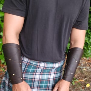 May include: Dark brown leather arm guards with gold-colored rivets and crisscross lacing. The arm guards are worn over a black t-shirt and a green, blue, and white plaid kilt. The arm guards are a medieval or fantasy accessory.