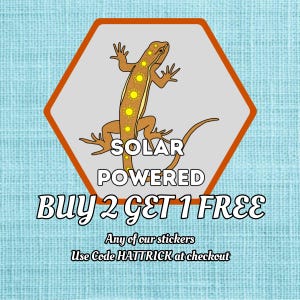May include: A sticker featuring a brown lizard with yellow sun designs, labeled "SOLAR POWERED." The sticker is in a gray hexagon with an orange border, and the text "BUY 2 GET 1 FREE" is below. The background is light blue.