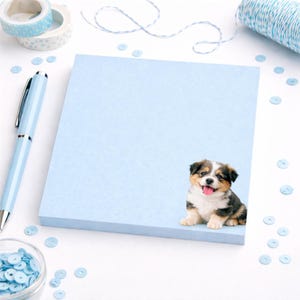 May include: A pale blue notepad featuring a puppy illustration. A blue pen, washi tape, and sequins are placed around the notepad. The puppy has brown, black, and white fur.