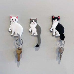 May include: Three cat-shaped key holders in white, gray, and black, each with a key ring attached. The white cat has pink ears, the gray cat has a gray tail, and the black cat has black and white markings.