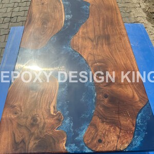 May include: A rectangular wooden table with a dark blue epoxy river design. The wood has a rich brown color with visible grain patterns. The text "EPOXY DESIGN KING" is visible on the table.