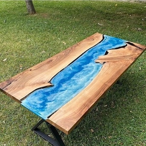 May include: A rectangular wooden table with a blue resin river running through the center. The table has a natural wood grain finish and sits on a black metal base. The blue resin has a cloudy, water-like appearance.