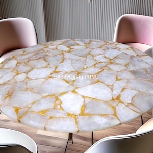 May include: A round table with a white and gold quartz-like surface. The table has a unique pattern with gold-colored veins running through the white stone. The table is supported by thin, black metal legs. The text "EpoxyDesignKING" is visible on the table.