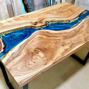 Puede incluir: Mesa de madera artesanal con un río de resina epoxi azul en el centro. La mesa tiene un acabado de veta de madera natural y descansa sobre patas de metal negro. El río de resina epoxi está acentuado con pequeñas piedras claras.