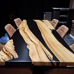 Puede incluir: Una mesa de comedor rectangular grande con un centro de resina epoxi negra y bordes de madera natural. La mesa está rodeada por seis sillas tapizadas marrones. La veta de la madera es visible y la mesa tiene una estética moderna y rústica.