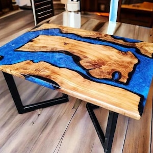 Pode incluir: Mesa retangular de madeira com um design de rio em resina epóxi azul. A mesa apresenta um padrão de grão de madeira natural com um acabamento brilhante, suportada por pernas de metal preto. A mesa tem aproximadamente 122 cm de comprimento.