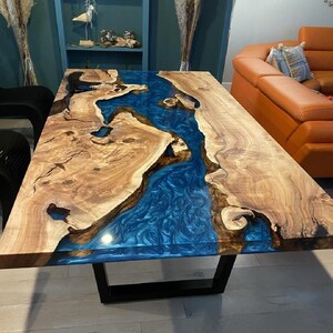 Op de afbeelding: Een handgemaakte houten tafel met een rivier van blauwe hars in het midden. De tafel heeft een natuurlijke houtnerf afwerking en een zwart metalen onderstel. De tafel is ongeveer 183 cm lang.