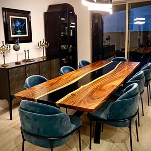 Op de afbeelding: Een grote houten eettafel met een zwarte epoxy rivier in het midden, omringd door acht teal fluwelen stoelen met zwarte metalen frames. De kamer heeft donkere houten meubels, kunstwerken en een moderne lamp.