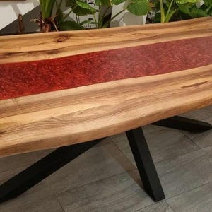 Pode incluir: Mesa de jantar de madeira com design de borda viva. O tampo apresenta uma incrustação central de resina vermelha, contrastando com o grão natural da madeira. A mesa é suportada por pernas de metal preto.