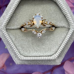 Può includere: Un set di anelli color oro con una pietra centrale di opale a forma di pera, accentuata da piccole pietre preziose trasparenti. La fascia ha un design delicato con dettagli a forma di luna crescente e foglie. Il set è esposto in un portagioie di velluto grigio.