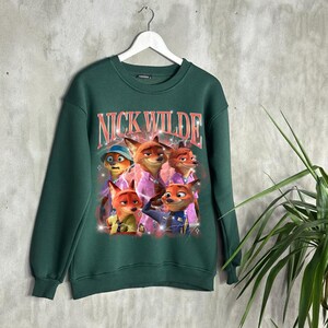 K&ouml;nnte beinhalten: Ein dunkelgr&uuml;nes Sweatshirt mit einer Grafik von Nick Wilde und anderen Charakteren aus Zootopia. Der Text "NICK WILDE" ist im Retro-Stil &uuml;ber den Charakteren aufgedruckt. Das Sweatshirt h&auml;ngt an einem wei&szlig;en Kleiderb&uuml;gel.