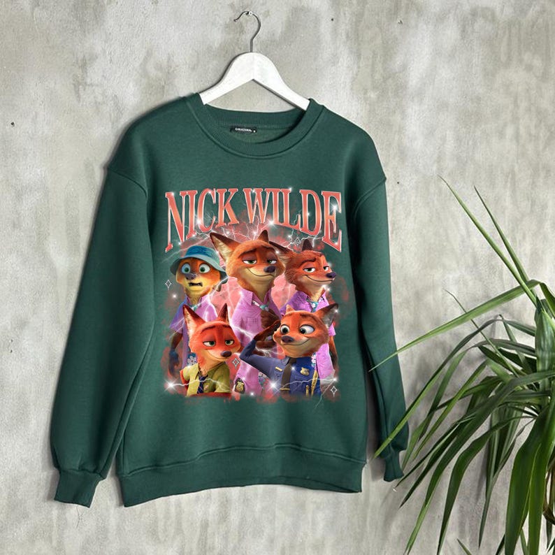 K&ouml;nnte beinhalten: Ein dunkelgr&uuml;nes Sweatshirt mit einer Grafik von Nick Wilde und anderen Charakteren aus Zootopia. Der Text "NICK WILDE" ist im Retro-Stil &uuml;ber den Charakteren aufgedruckt. Das Sweatshirt h&auml;ngt an einem wei&szlig;en Kleiderb&uuml;gel.