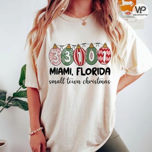 Może przedstawiać: Kremowa koszulka z świątecznym wzorem. Grafika zawiera liczby 33101 w stylu bombek choinkowych, nad słowami "MIAMI, FLORIDA" i "small town christmas" w czarnym tekście.