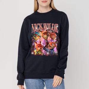 K&ouml;nnte beinhalten: Marineblaues Sweatshirt mit einem Aufdruck der Figur Nick Wilde aus dem Animationsfilm Zootopia. Das Bild enth&auml;lt den Text "NICK WILDE" in einer Retro-Schriftart, mit mehreren Cartoon-Fuchs-Charakteren. Das Sweatshirt hat lange &Auml;rmel.