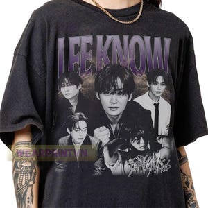 May include: Dark gray t-shirt featuring a black and white image of Lee Know from Stray Kids. The shirt has the name "LEE KNOW" in purple block letters across the top. The band's name, "Stray Kids", is printed in white script at the bottom.