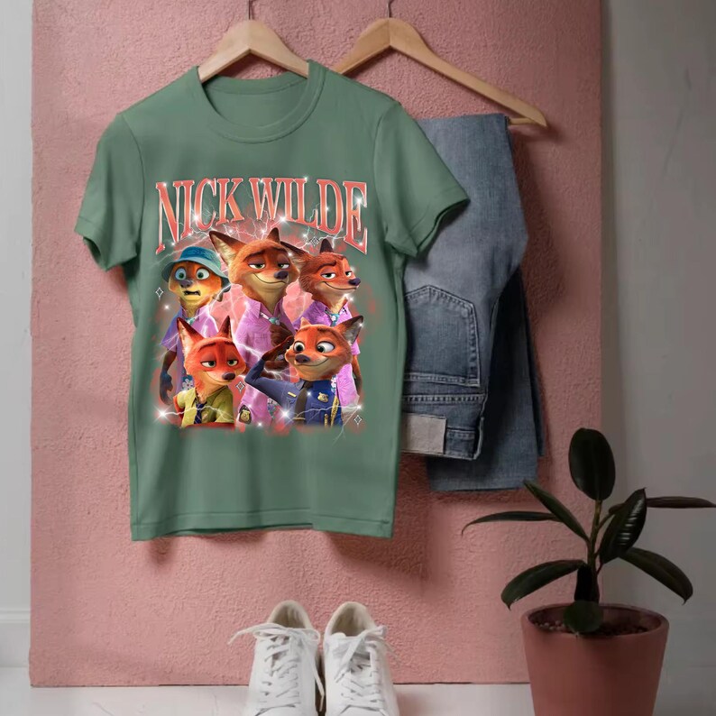 K&ouml;nnte beinhalten: Salbeigr&uuml;nes T-Shirt mit einer Grafik von Nick Wilde aus Zootopia. Die Grafik enth&auml;lt mehrere Bilder von Nick Wilde mit dem Text "NICK WILDE" in einer Retro-Schriftart. Das Shirt h&auml;ngt mit blauen Jeans und wei&szlig;en Turnschuhen.