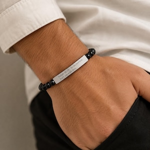 Könnte beinhalten: Ein Armband aus glatten, runden schwarzen Perlen und einer rechteckigen silberfarbenen Metallplatte. Die Platte ist mit dem Text graviert: Isaiah 41:10 - So do not fear, for I am with you. Die Perlen sind auf einem flexiblen Band aufgezogen.