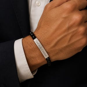 May include: A beaded bracelet featuring smooth, round black beads and a rectangular silver-toned metal bar. The metal bar is engraved with the text "God is in Control" in a clear, dark font. The beads are strung together in a continuous loop.